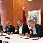 four men in suits, seated at a table with one man signing a document while the others watch