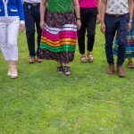 Seven people walking together, shown from the waist down, with two wearing ribbon skirts and one in a Métis sash.