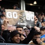 Group of fans cheering on the Bison football team and holding a sign that says 'Go Bisons'.