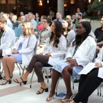 Five MPAS students wearing white coats and stethoscopes are sitting in chairs. A crowd of people is sitting behind them.