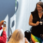 Two people pose while a man takes a photo of them with his cell phone.