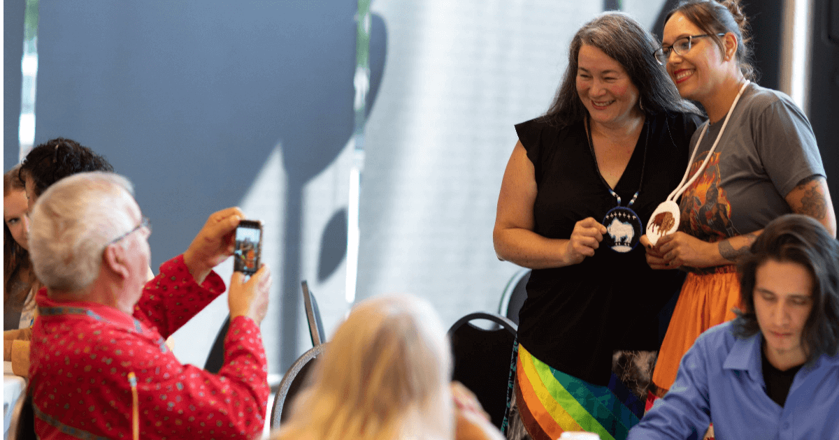 Two people pose while a man takes a photo of them with his cell phone.