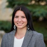 An Indigenous woman in a grey blazer and beaded earrings smiles at the camera.