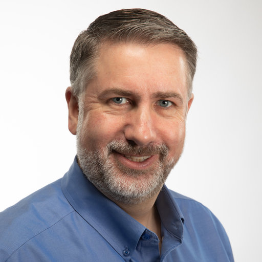 Man wearing a blue collared shirt with salt and pepper hair and short beard.