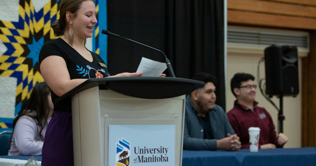 A woman speaking at a podium with a star blanket behind her and a panel of speakers beside her, sitting at a table.