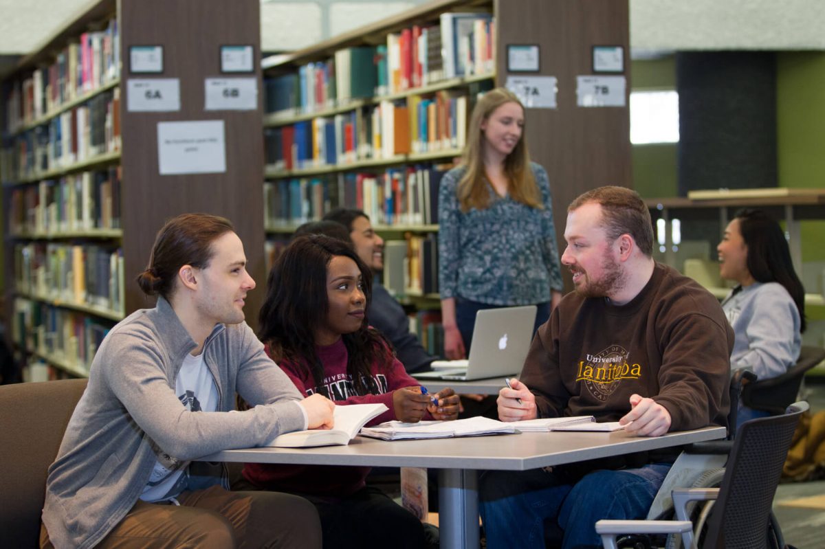 Three students sit around a round study table surrounded by shelves of library books.