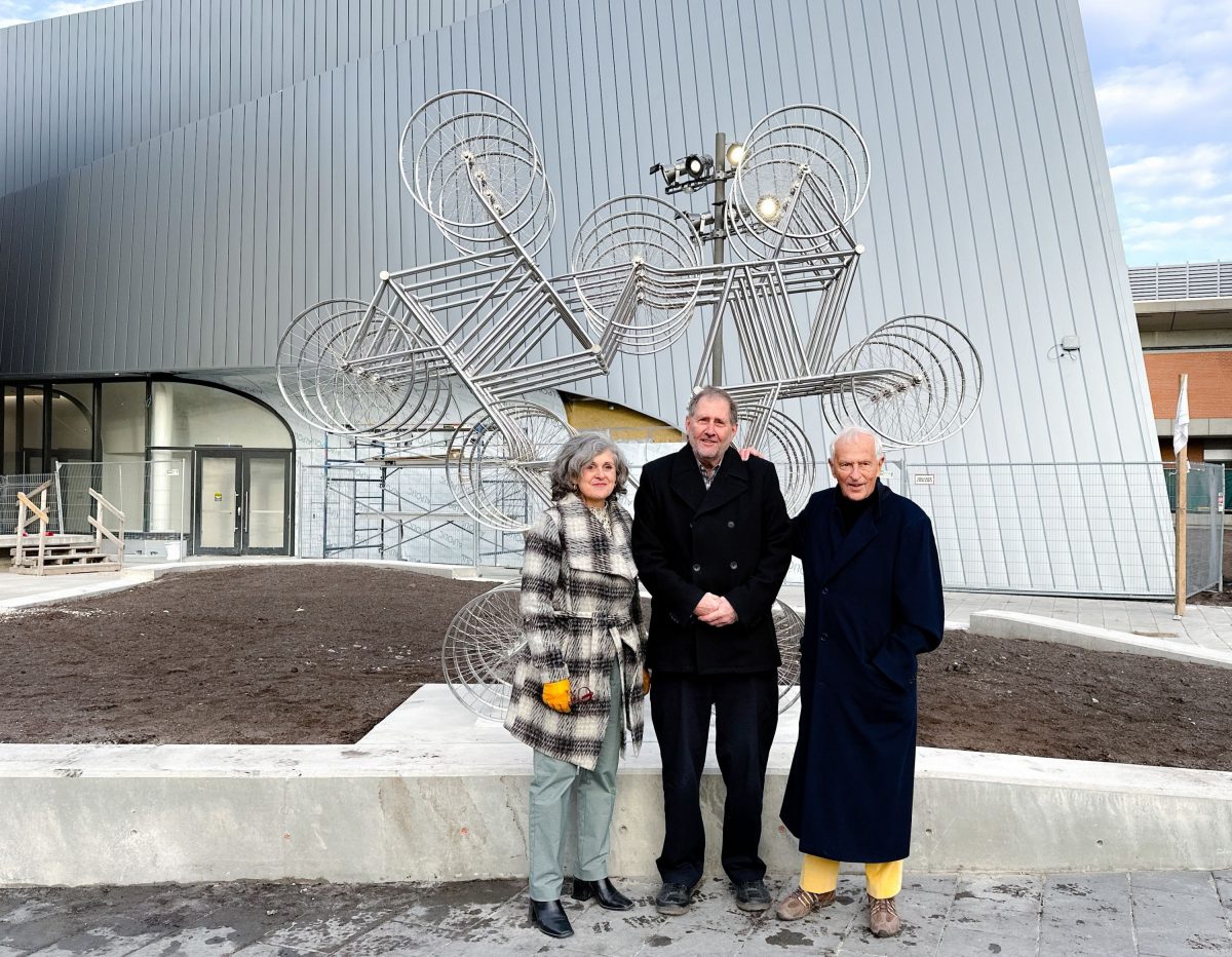 Three people stand in front of a bicycle sculpture made of 18 bicycles. The Desautels Concert Hall is in the background.
