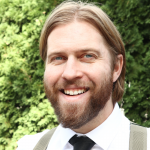 portrait of Jordan Laidlaw. He is wearing a dress shirt, necktie, and has a beard and mustache. He is smiling and standing outdoors.