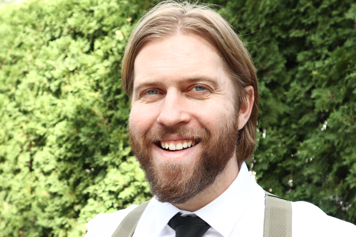 portrait of Jordan Laidlaw. He is wearing a dress shirt, necktie, and has a beard and mustache. He is smiling and standing outdoors.