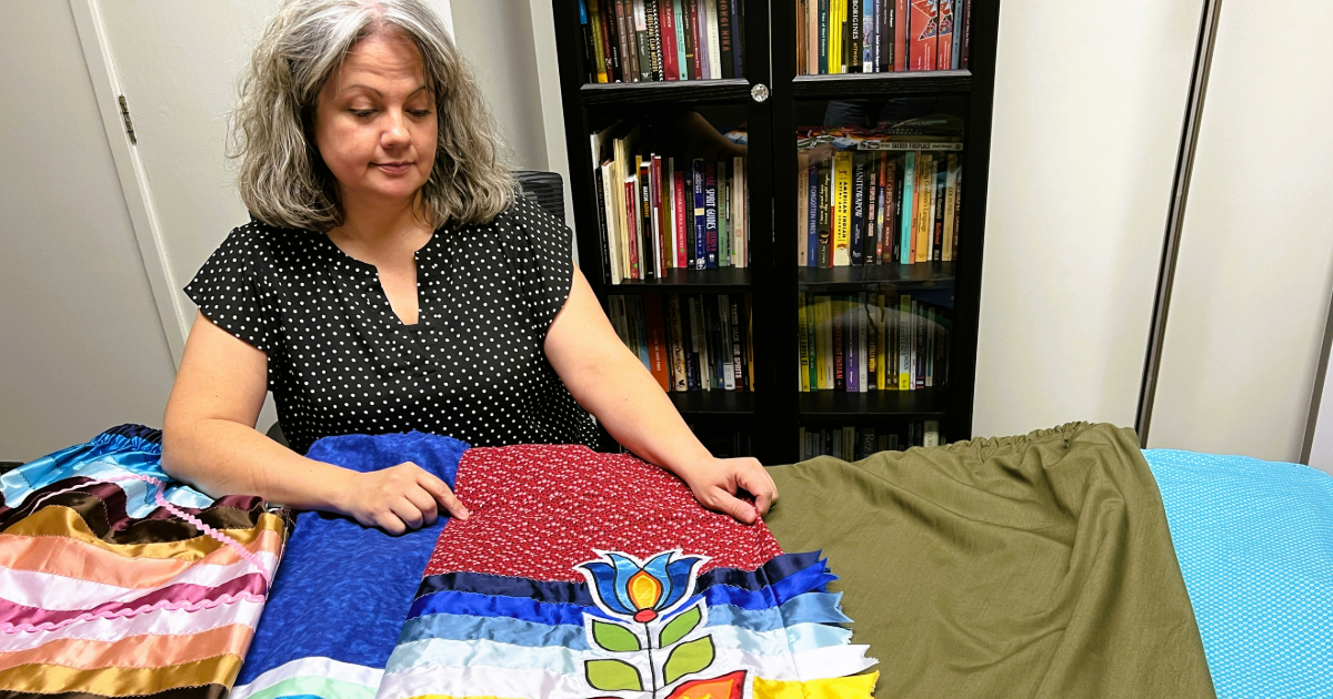 An Indigenous woman sits behind a table of colourful ribbon skirts.