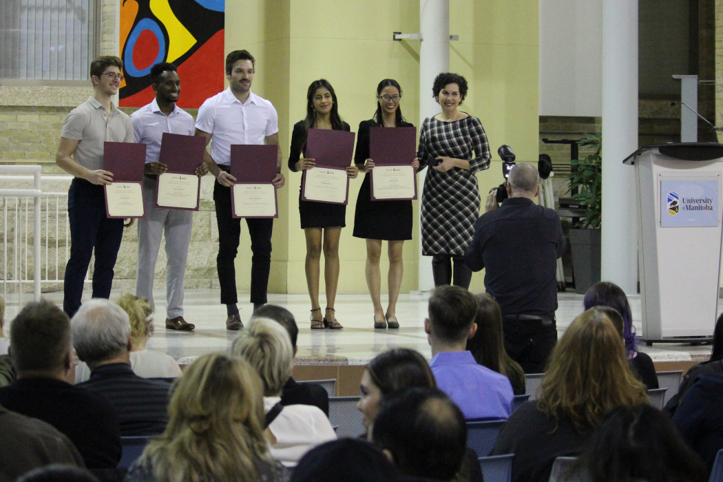 Five students stand in a line with Dr. Kelekis-Cholakis on a stage. A photographer is taking their photo. The students are holding certificates.