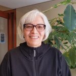 Photo of Wendy Whitecloud wearing a black hoodie with the hood down, standing in front of a large green plant with big leaves in the law library. A door to another room behind her stands open.