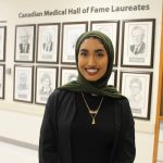 Mirha Zohair poses in front of 13 framed illustrations of medical hall of fame laureates from UM. Text on wall reads "Canadian Medical Hall of Fame Laureates."