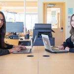 Two Indigenous students sitting in a computer lab, both facing the camera and smiling.