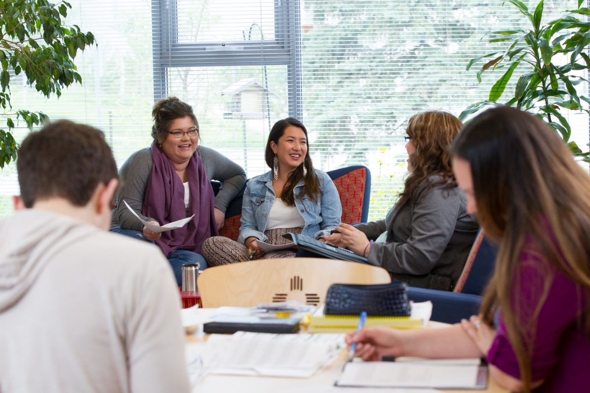 A group of Indigenous students gather around a table. Several are talking with each other and smiling, while others write in notebooks.