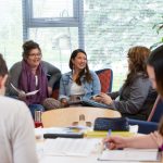 A group of Indigenous students gather around a table. Several are talking with each other and smiling, while others write in notebooks.