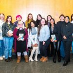 Group of 15 students standing in front of a wooden wall, most holding a book.