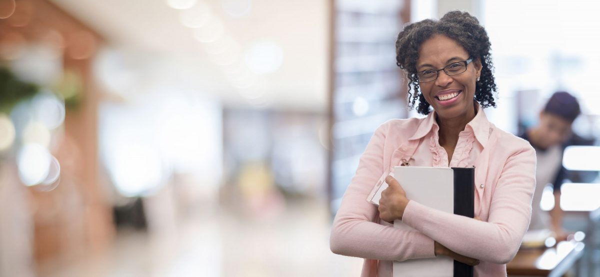 Woman wear a pink shirt smiling and holding a book in a hallway.