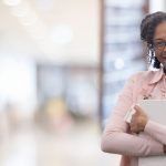 Woman wear a pink shirt smiling and holding a book in a hallway.
