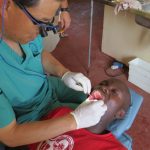 Patient in a dental chair with their mouth open. Dr. Kim performs dental treatment while wearing white gloves and holding dental instruments.
