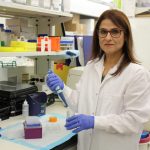 Dr. Soheila Karimi holds a pipette and a jar. She is in her lab and is wearing gloves and a lab coat.