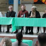 Five physicians sit behind two tables on a stage. One of them is speaking into a microphone.
