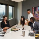 Five students working in a boardroom with the city of Winnipeg skyline in the bakground.
