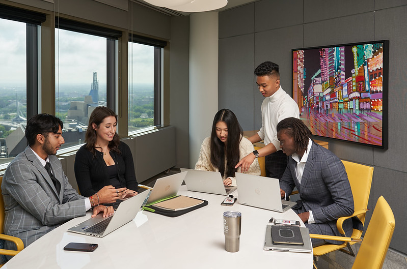 Five students working in a boardroom with the city of Winnipeg skyline in the bakground.
