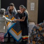 A man is wrapped in a star blanket by two Elders at a recognition ceremony.