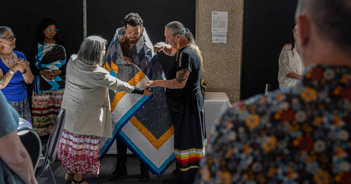 A man is wrapped in a star blanket by two Elders at a recognition ceremony.