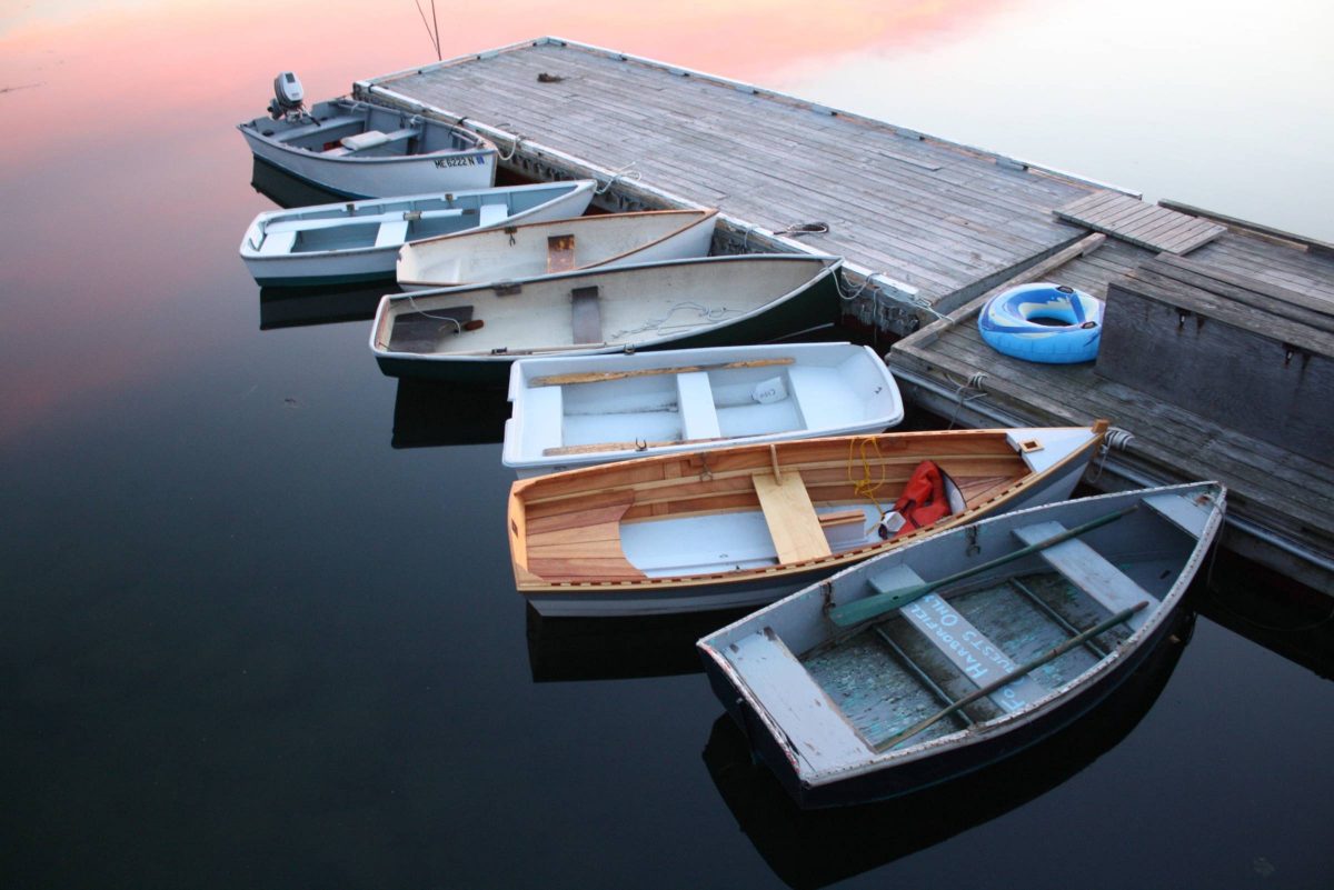 Seven two-man fishing boats, of a variety of sizes, styles, materials, and ages sit tied up to a weathered wood dock on a perfectly calm lake during sunset.