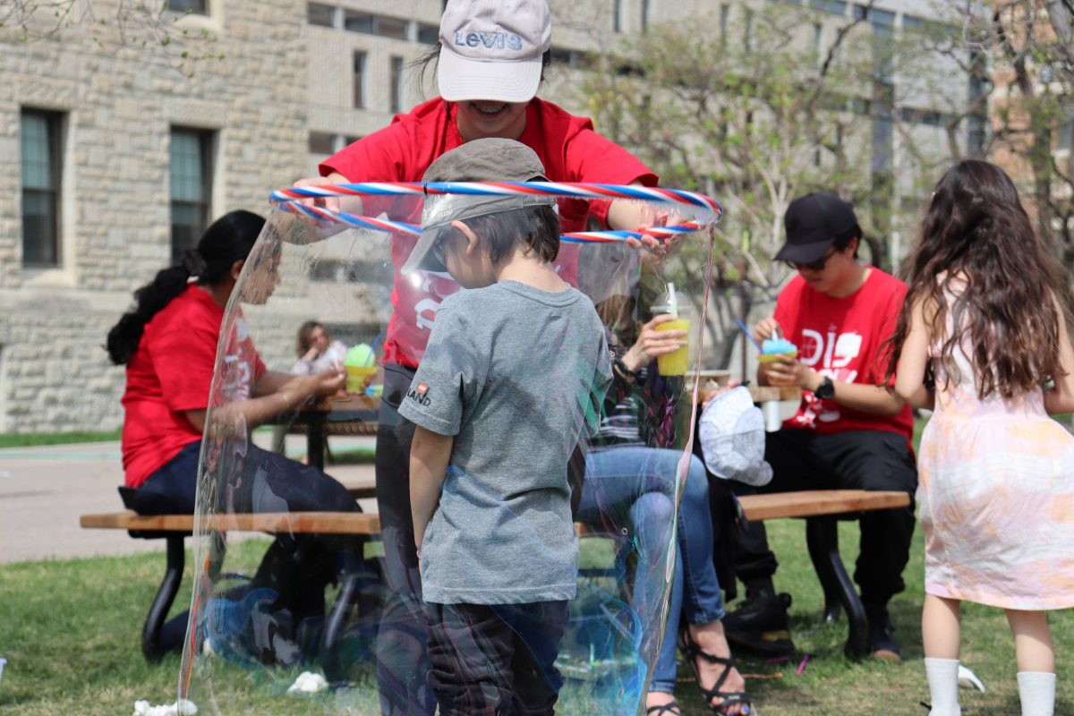 A kid with a grey t-shirt and cap standing in the middle of a large bubble created around him by a student wearing a red shirt with soap and hula loop in front of the science buildings complex outside on the grass with kids and other people in the background.