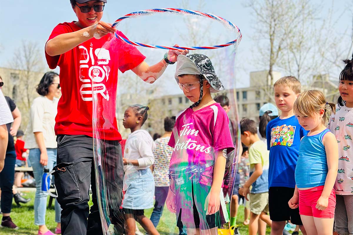 A kid in the middle of a bubble made with soap and hula loop by a university student wearing a red Science Rendezvous volunteer shirt.