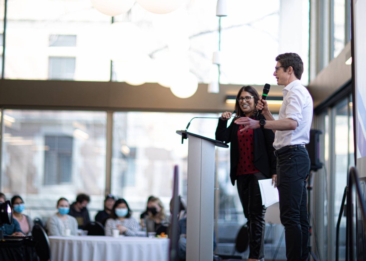 Wide shot of the profile a young man with white shirt and black pants on the stage talking with a wireless microphone facing the audience with a woman standing next to him smiling behind a podium.