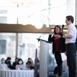 Wide shot of the profile a young man with white shirt and black pants on the stage talking with a wireless microphone facing the audience with a woman standing next to him smiling behind a podium.
