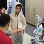 A boy around 12 years old checks an adult's blood pressure in a classroom setting.