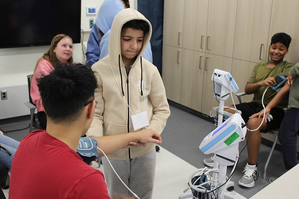 A boy around 12 years old checks an adult's blood pressure in a classroom setting.