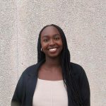 Witta Irumva, Black female student standing on front of a grey wall, wearing a black and white outfit, smiling.