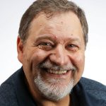 Man with short brown hair, grey short beard, big smile and a dark blue collared shirt.