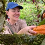 A young woman picking a cacao pod from a plant in rainforest setting.