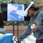 A woman wearing a VR headset and hand controls in front of a TV showing a virtual hospital room.