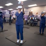 Nursing students in a lab setting wearing scrubs and virtual reality headsets and hand controls reach as they work in virtual environments. Other students in the background look at computer screens.