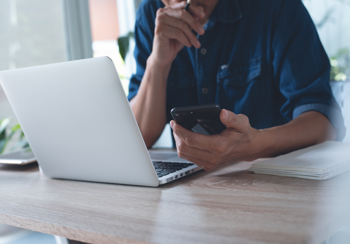 A person working on a laptop computer and holding a cellphone in their left hand.