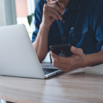 A person working on a laptop computer and holding a cellphone in their left hand.