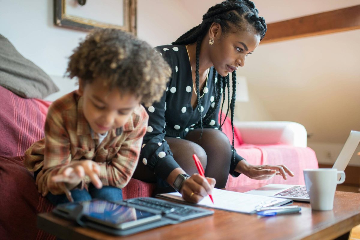 Racialized mother working from home on a couch staring at a computer, with child out of focus playing on a tablet.