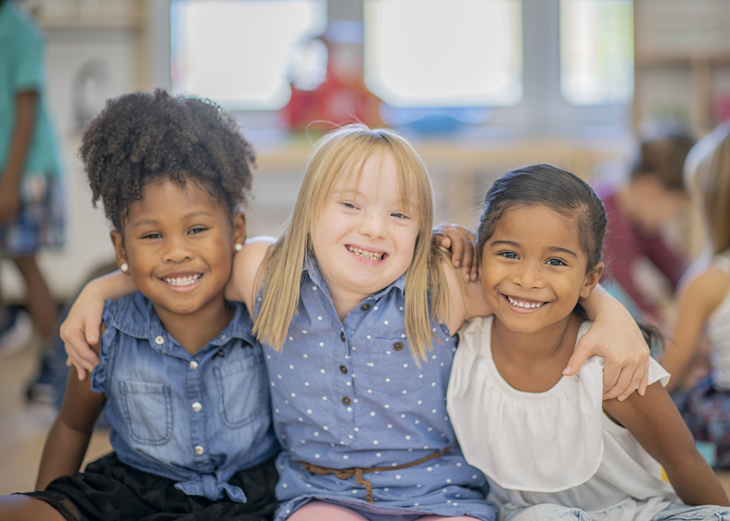 Three little girls of different ethnicities and abilities smile and put their arms around each other in a kindergarten classroom.