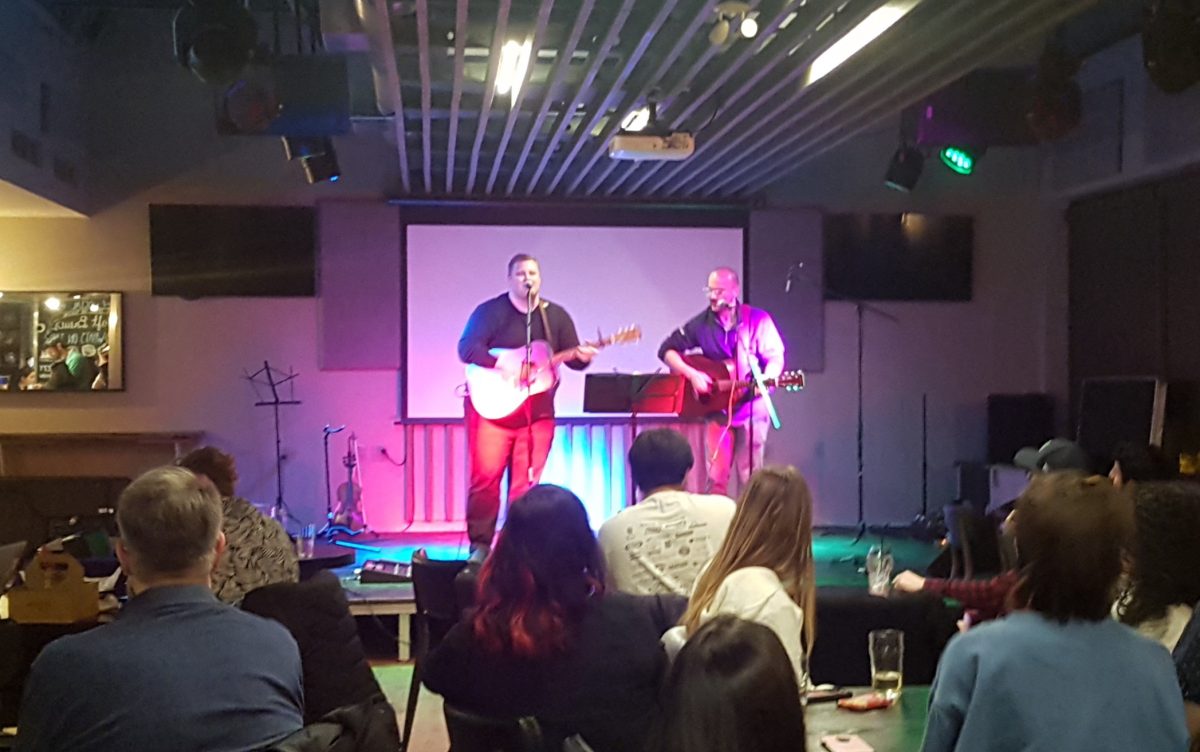 Two young men playing guitar and singing on stage while an audience looks on.