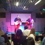 Two young men playing guitar and singing on stage while an audience looks on.