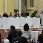 The five panelists sit at a table on a stage. One of the panelists is talking into a microphone. The moderator stands behind a lectern.