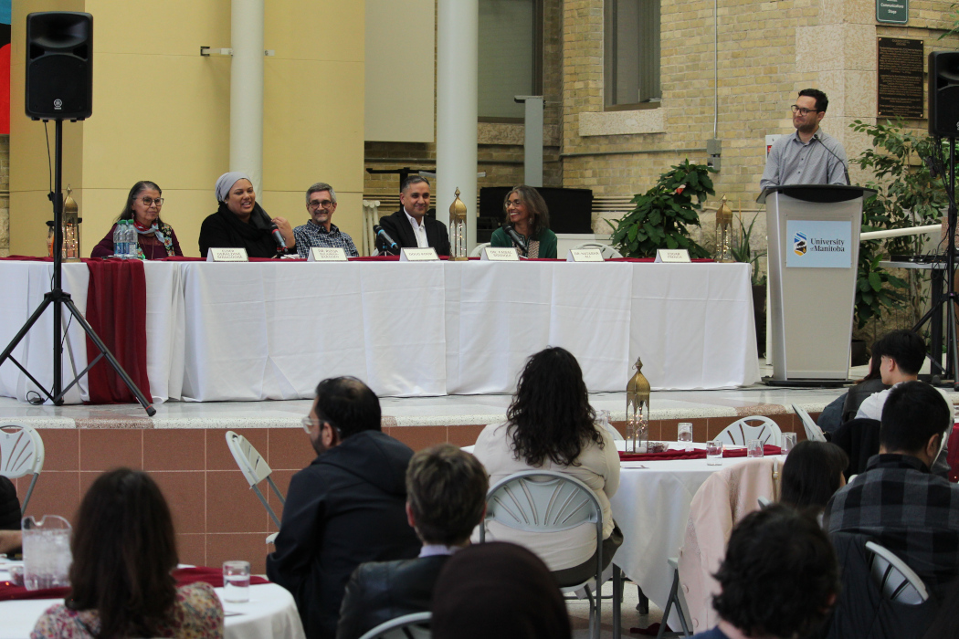 The five panelists sit at a table on a stage. One of the panelists is talking into a microphone. The moderator stands behind a lectern.